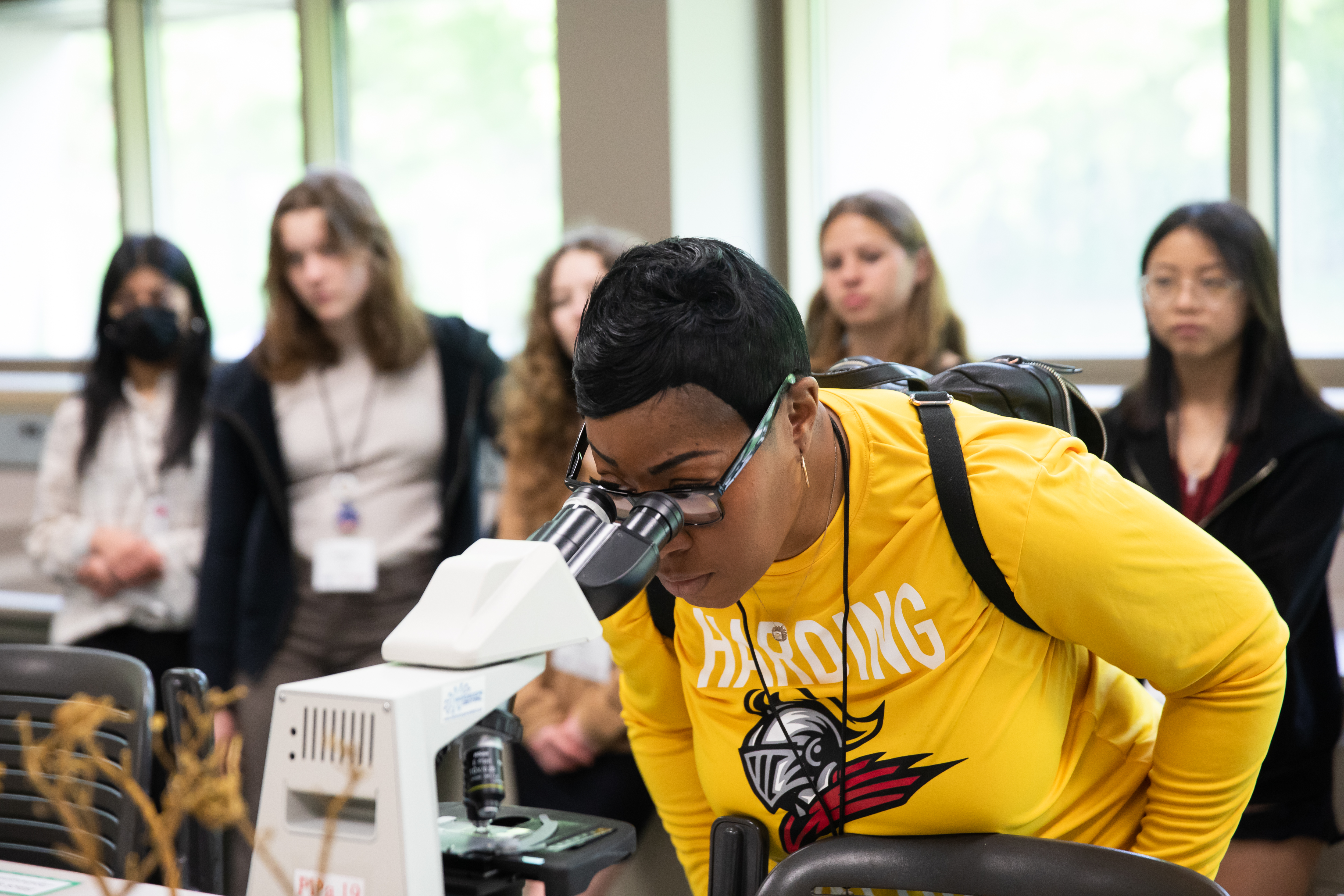 A Black woman in a yellow shirt peers into a microscope with a row of students lined behind observing